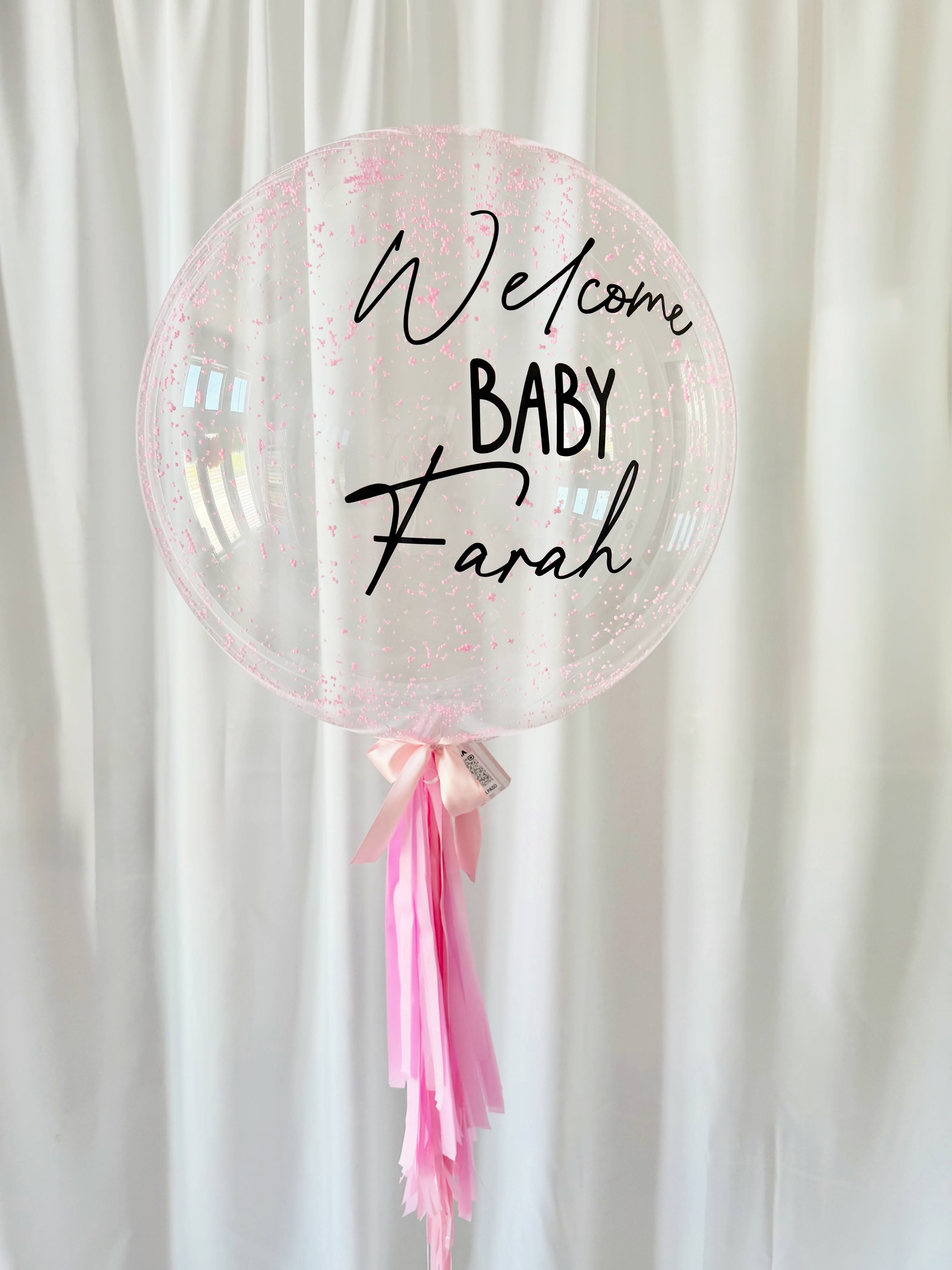 Globo de bienvenida a un bebé con pequeñas motas rosas, letras negras y cintas rosas.