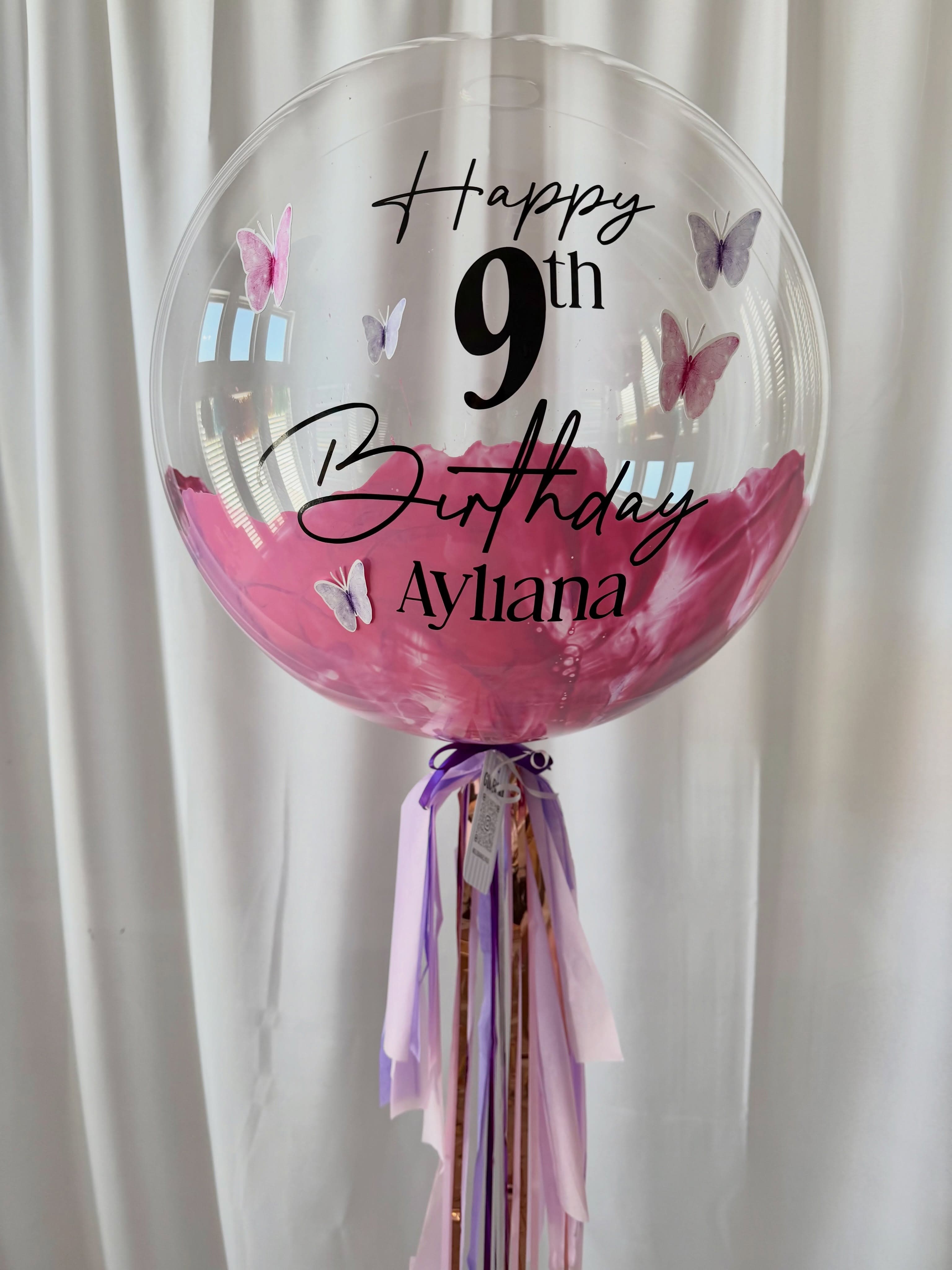 Globo de cumpleaños con mariposas rosas y lilas, letras negras y cintas de colores.